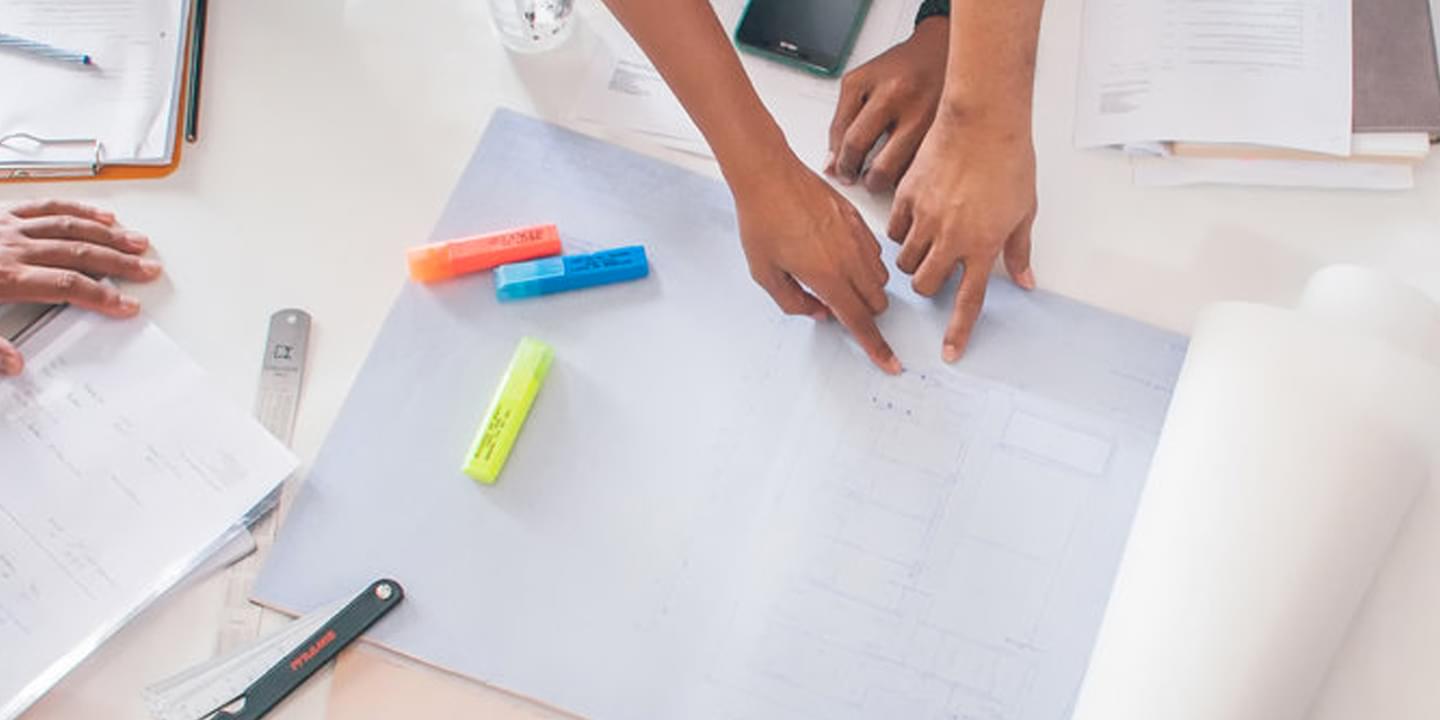 Overhead shot of a desk covered in various papers, writing instruments, and clipboards. Hands belong to people of various ages, races, and genders reach in from off screen to point at different pages.