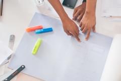 Overhead shot of a desk covered in various papers, writing instruments, and clipboards. Hands belong to people of various ages, races, and genders reach in from off screen to point at different pages.