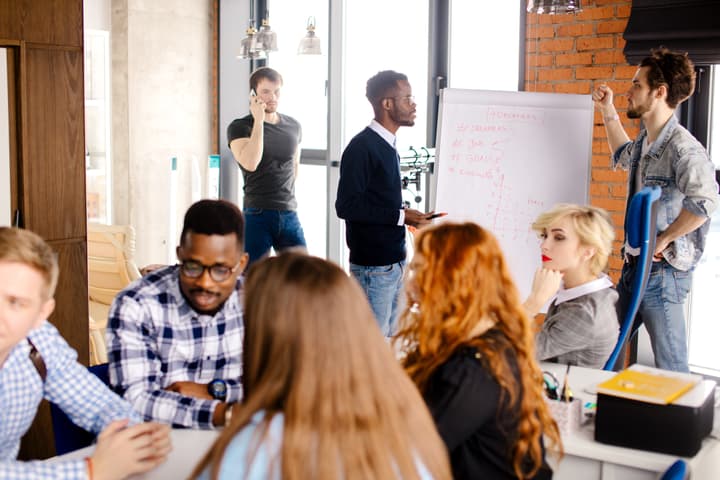 Seven business people in casual dress of various races and genders collaborate in smaller groups around a modern office space.