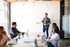 A white business man in casual attire stands in front of a large whiteboard and presents to a modern meeting room to colleagues of various ages, races, and genders.
