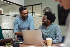One male stands while talking to another male sitting at a table with a laptop having a friendly discussion while smiling.