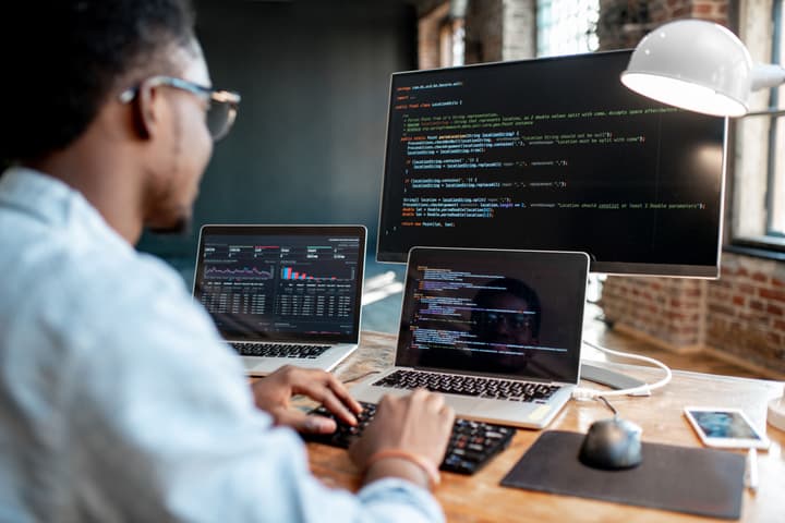 A Black Engineer sitting in front of computer with multiple screens coding
