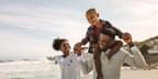 A Black family of three, comprised of a mother, father, and young boy, take a vacation on the beach. The young boy sits on the father's shoulders. He holds hands with both his father and mother.