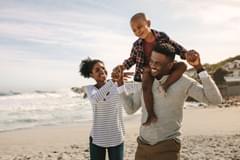 A Black family of three, comprised of a mother, father, and young boy, take a vacation on the beach. The young boy sits on the father's shoulders. He holds hands with both his father and mother.