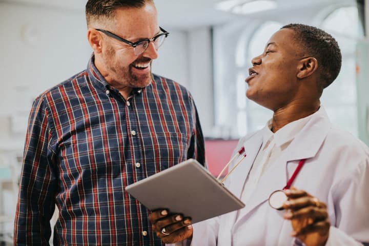 A white man in plain clothes talks to a Black man wearing a lab coat and a stethoscope. They are having a lively and friendly conversation. The Black man holds a digital tablet.