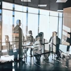 Conference being held in a modern office with glass walls. Several people inside are obscured by glares on the glass from the sun.