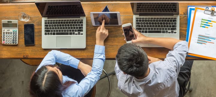 Two business people discuss sales figures while seated at a high bar that has a calculator, two smart phones, two laptops, and clipboards with documents.