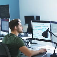 A young, white male in casual clothes works in a modern office with lots of computers and computer monitors. He is focusing on two monitors in front of him filled with lines of code.