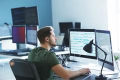 A young, white male in casual clothes works in a modern office with lots of computers and computer monitors. He is focusing on two monitors in front of him filled with lines of code.