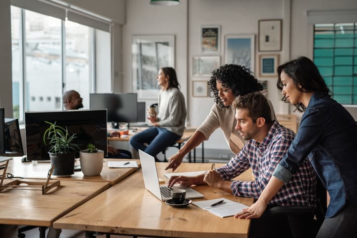 Man sits while two female colleagues stand around him while they all look at a laptop on a desk.