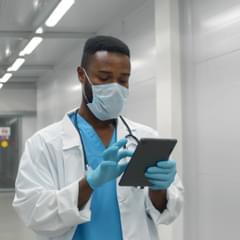 A Black male doctor wearing protective mask and gloves using tablet working in medical clinic.