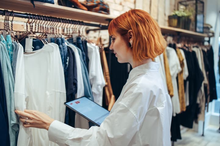 A white woman takes inventory in a retail clothing store with an electronic tablet.