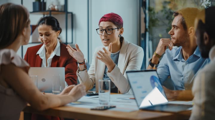 A diverse group of people sit around a conference room table with laptops having a business meeting