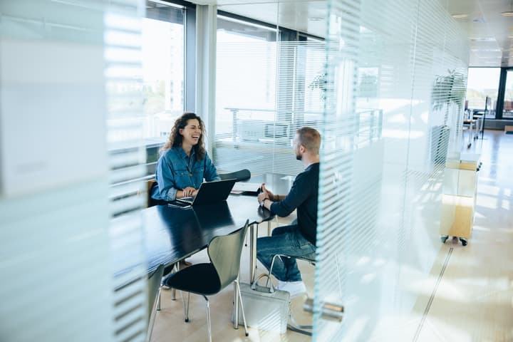 A woman sits at a table in front of a computer talking with a man.