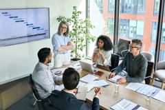 Shot from above into conference room where a white older woman is standing and four people are sitting and having a strategic discussion