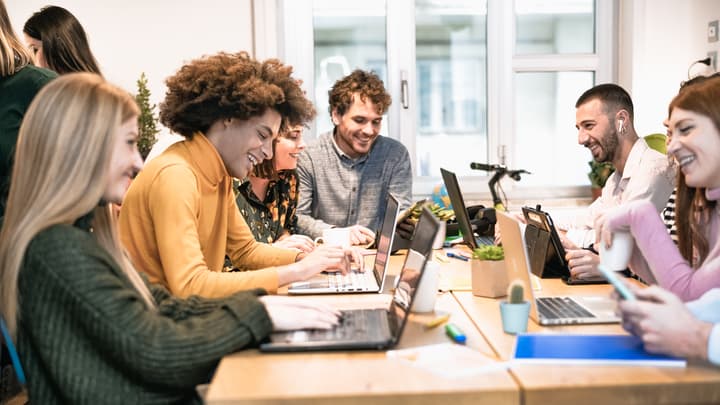 Young business people of various races and genders are gathered around a coworking table with laptops. They appear to be having a fun and lively conversation.