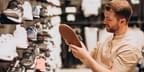 A young caucasian man with facial hair chooses a pair of sneakers from a wall of shoes in a footwear store.
