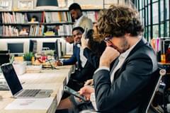 White businessman with curly hair wears a business suit and has his head on his chin as if deep in thought as he looks at this mobile phone. Three other coworkers of various ages, genders, and races are in the background and appear to be conversing at the meeting table they are all seated at.