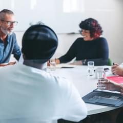 A diverse group of six businesspeople of various ages, races, and genders gathered around a circular meeting table in a modern office. They appear to be having an energetic and positive discussion.