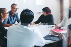 A diverse group of six businesspeople of various ages, races, and genders gathered around a circular meeting table in a modern office. They appear to be having an energetic and positive discussion.