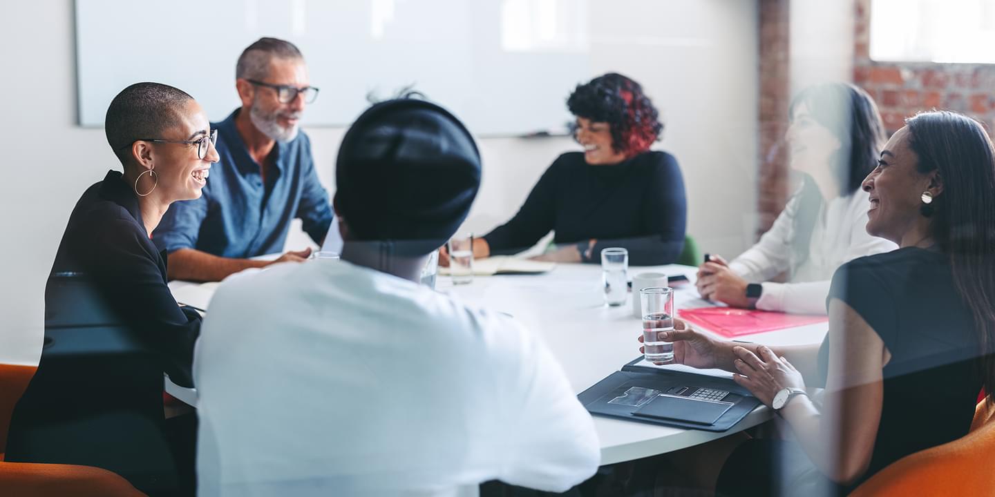 A diverse group of six businesspeople of various ages, races, and genders gathered around a circular meeting table in a modern office. They appear to be having an energetic and positive discussion.