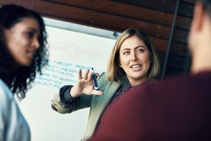 A white businesswoman in business casual attire explains a concept to her colleagues.