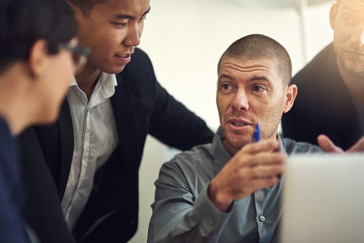 A middle-aged man seriously explains something to his colleagues while seated in front of a laptop computer.