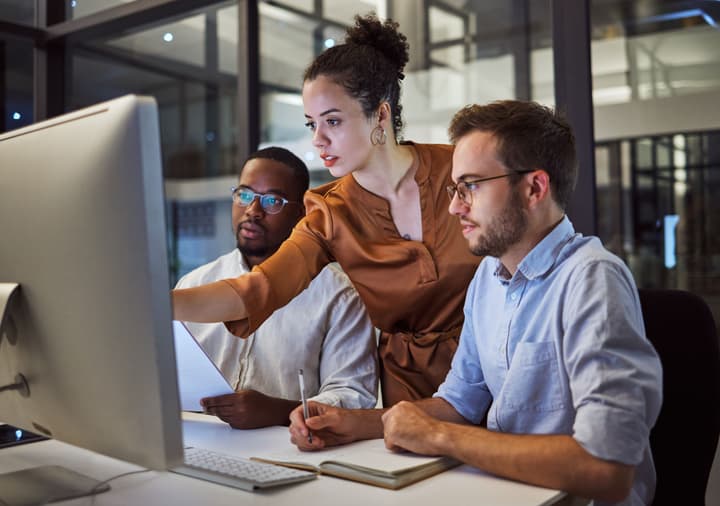 Woman of color speaking to two men sitting while pointing at a computer monitor in an office discussing technology planning ideas and KPI strategy.