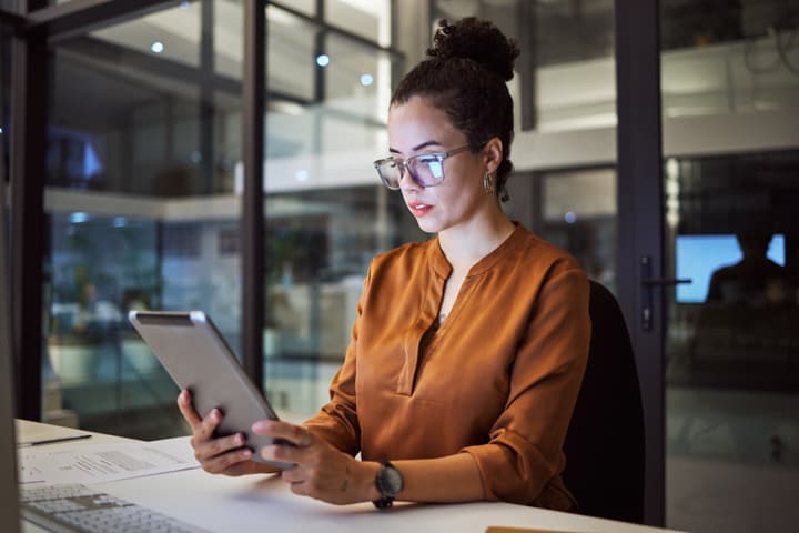 A white businesswoman working in an office at night, looking at a tablet.