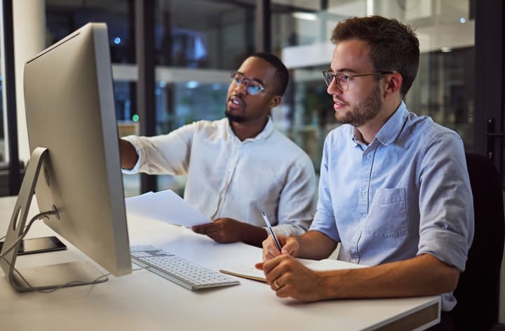 Two male colleagues, one Black, the other white, work together on a desktop computer analyzing data.