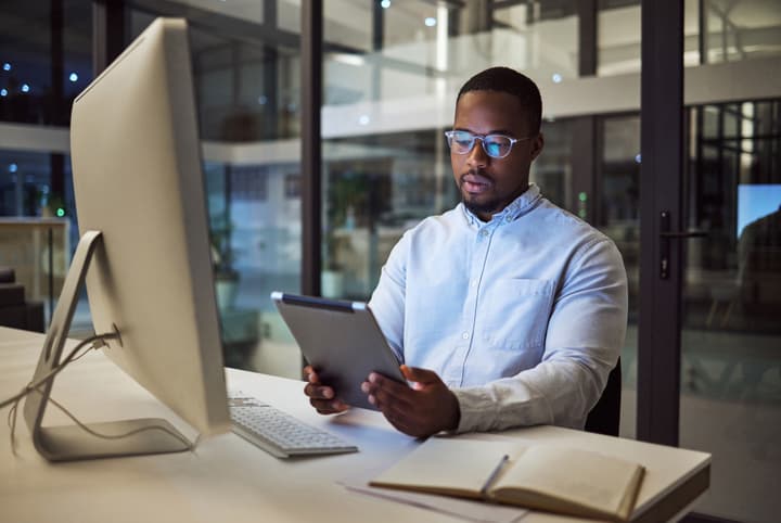 A young black businessman sits in front of a computer holding a tablet in an office building in the evening.