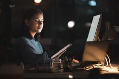 A woman of color works in an office at night. Her face is illuminated by the glow of her computer monitors. She holds a tablet in one hand, and is checking a laptop with the other hand. There is a large external monitor on her desk as well.