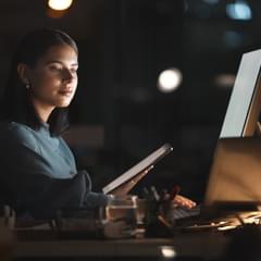 A woman of color works in an office at night. Her face is illuminated by the glow of her computer monitors. She holds a tablet in one hand, and is checking a laptop with the other hand. There is a large external monitor on her desk as well.