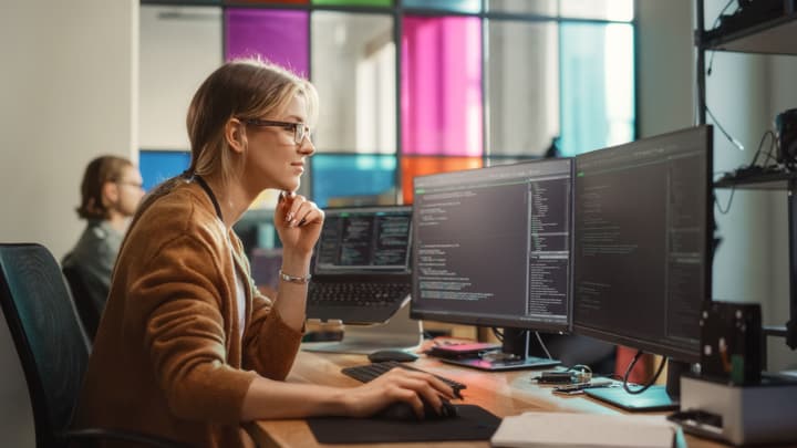 Caucasian Female Software Engineer Writing Code on Desktop Computer with Multiple Screens Setup in Office Space.