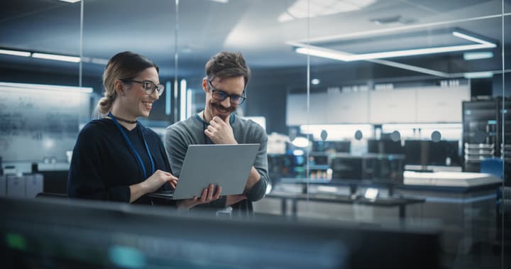 A woman is holding a laptop while a man looks at the screen smiling while in an office