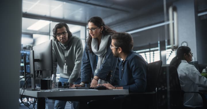 Two people are standing and leaning on a desk while a third person sits at the desk with a computer as they talk
