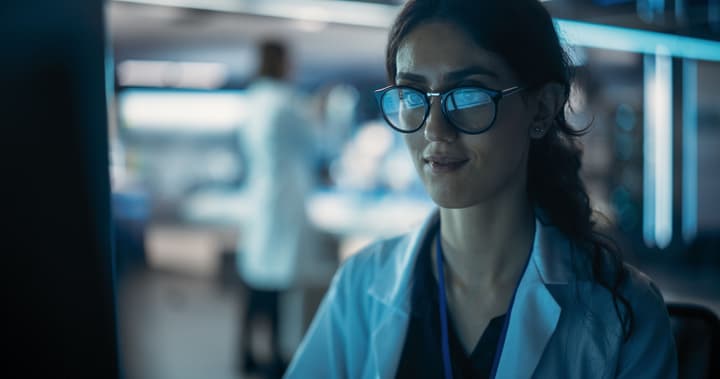 A female doctor with glasses looks at a computer screen with a blurred background