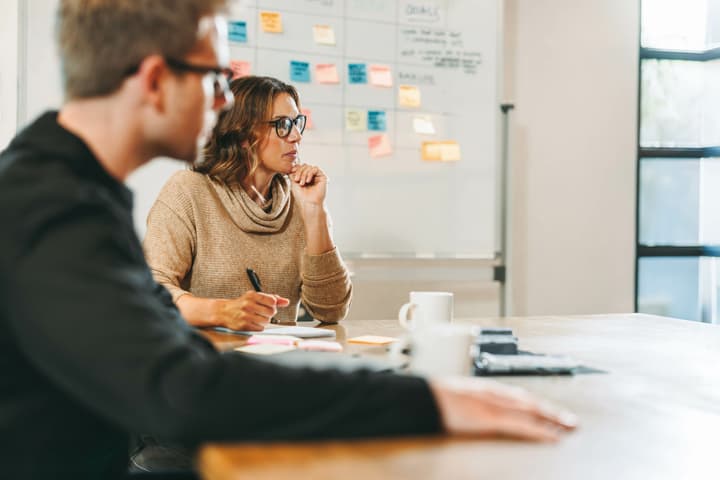 A woman sits at a conference table taking notes with a man in the foreground and a dry erase board with sticky notes in the background