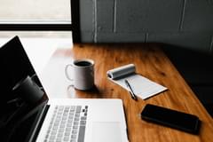 A wooden desk with the following resting on its surface: a MacBook laptop, a memo pad with illegible writing, a white mug full of black coffee, and a smart phone. Photo taken by Andrew Neel via Unsplash