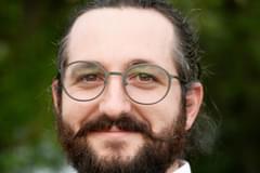 A portrait of Propeller consultant Alexander Winston. He wears round, black-rimmed glasses, a dark blue blazer, and a white button-up shirt.
