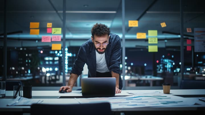 Businessman with glasses and facial hair Working on Laptop Computer in Big City Office Conference Room Late in the Evening