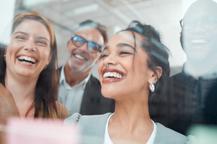 Four coworkers smiling and laughing in front of a glass wall with sticky notes on the edges.