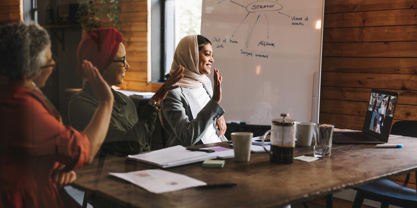 Female colleagues sitting a conference room table with a whiteboard in the background smiling and waving at people on a laptop in a virtual meeting