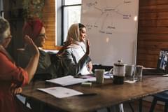 Female colleagues sitting a conference room table with a whiteboard in the background smiling and waving at people on a laptop in a virtual meeting