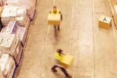Overhead photograph of two workers in yellow, hi-vis vests moving large, cardboard boxes around a stock warehouse. They are moving so quickly both figures are blurred.