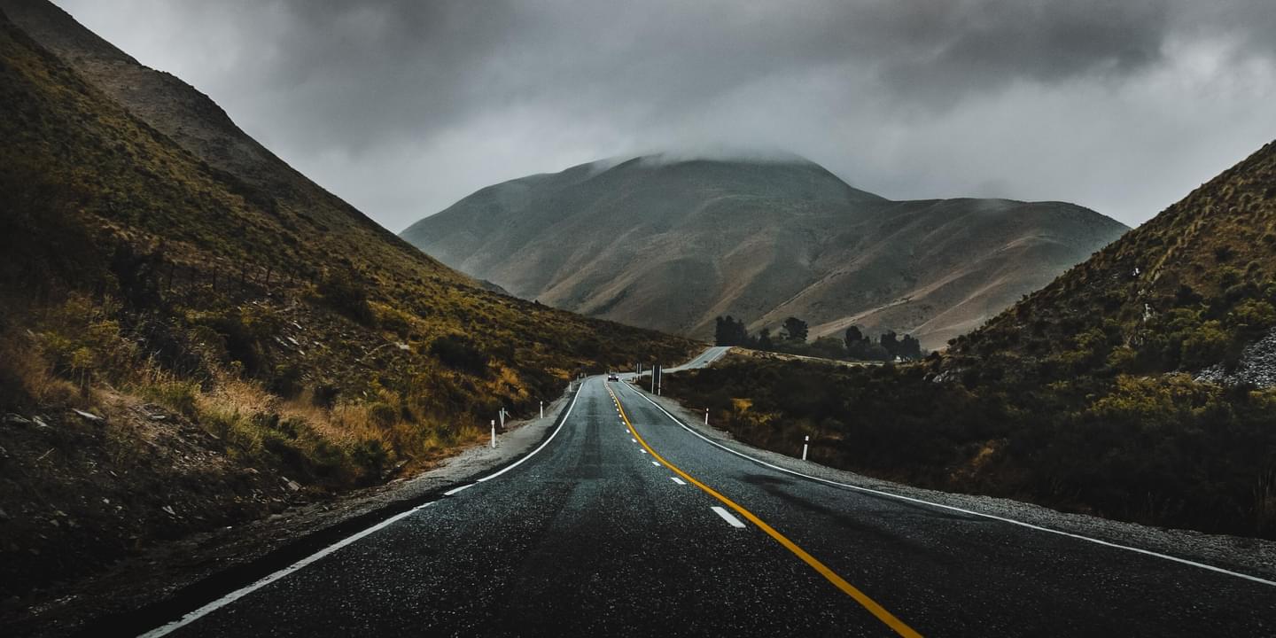 A paved, two lane road winding through desert hills on a gloomy, overcast day. Image by Iswanto Arif via Unsplash.