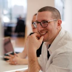 A millennial-aged white male smiles in a modern office setting. He appears to be engaged in a lively conversation with colleagues.