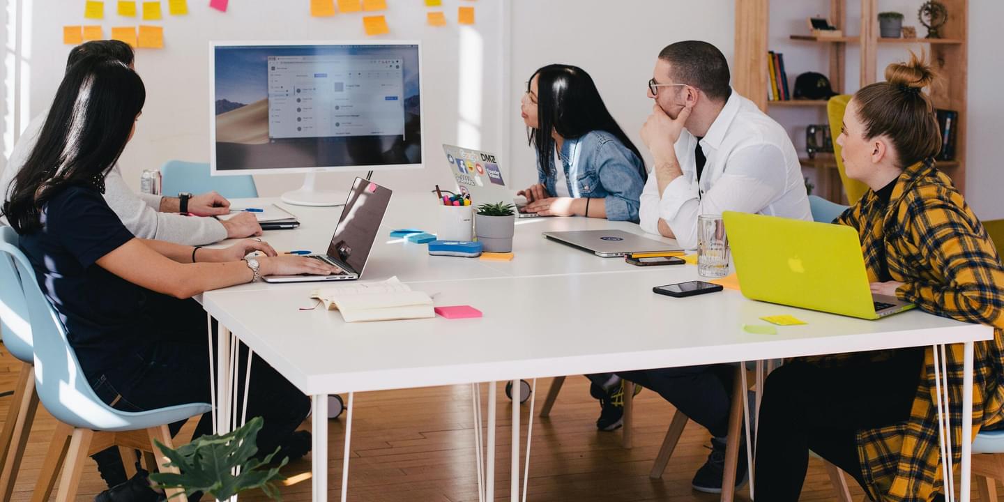 A small group of millennial-aged professionals of various races and genders gather around a modern, bright meeting space with their laptops to work on a project. Photo by Jason Goodman via Unsplash.