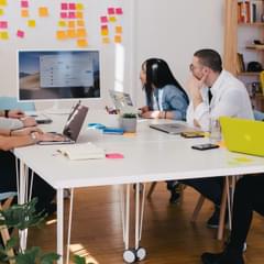 A small group of millennial-aged professionals of various races and genders gather around a modern, bright meeting space with their laptops to work on a project. Photo by Jason Goodman via Unsplash.