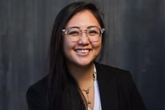 A portrait of Propeller employee Lena Chhay. She wears a white blouse and a black blazer in front of a dark, grey, wooden background. She wears glasses with a clear, acrylic frame.